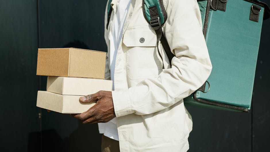 A person holding three cardboard boxes stacked on top of each other, with the top box being smaller and light brown, while the lower boxes are larger and beige. The individual is dressed in a light-co