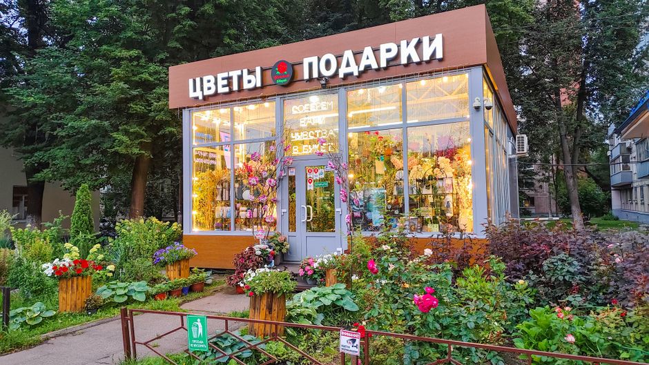 Exterior of a flower shop with large glass windows displaying vibrant floral arrangements inside, illuminated by warm lighting. The shop has a modern, wooden facade with a sign in Russian indicating '