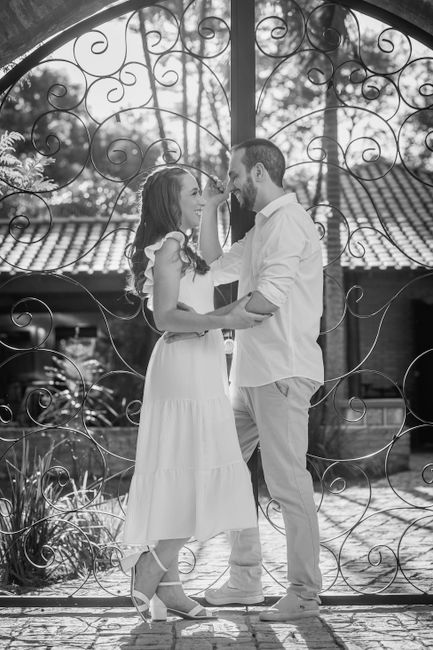A black-and-white photograph of a couple standing close together beneath an ornate, wrought-iron gate with intricate swirling patterns. The woman wears a flowing, ankle-length white dress with ruffled