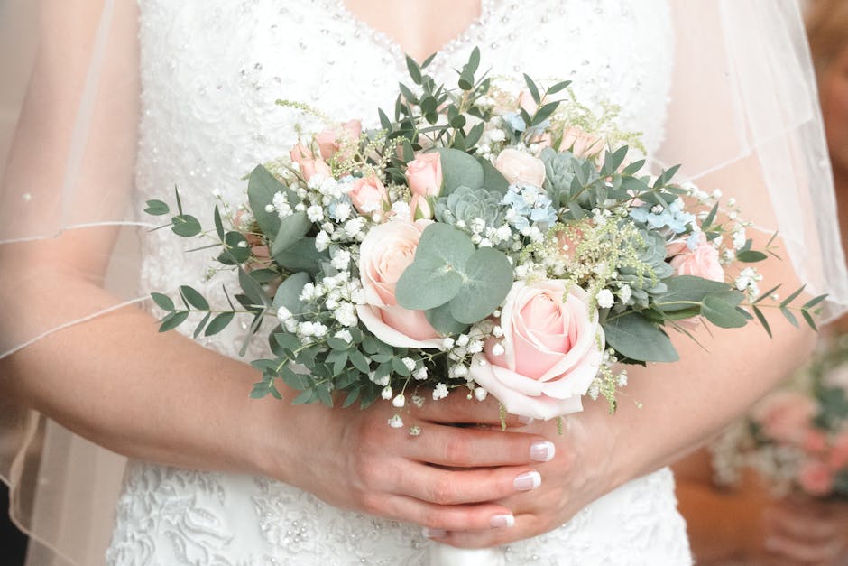 A bride is holding a delicate floral bouquet featuring pale pink roses, sprigs of white baby's breath, eucalyptus leaves, and soft blue-green succulents. The bouquet is wrapped in subtle white paper a