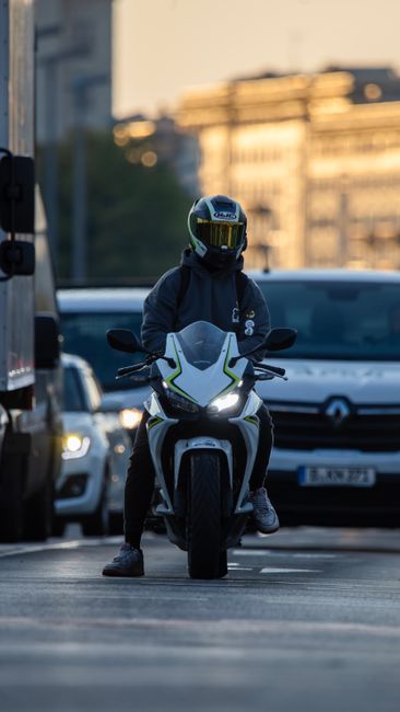 A person wearing a dark blue jacket and a full-face helmet with a reflective visor is sitting on a white and black motorcycle, positioned on a city street during daylight. Behind them, there are multi