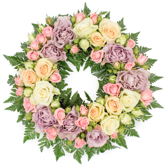 A beautifully crafted funeral wreath sits perfectly centred against a plain white background, its circular form completely covered with roses and lush greenery so that the eye travels smoothly around the ring. The design looks like it has been made by an experienced florist serving families in Chadwell Heath, with each stem carefully placed to create balance and harmony. Large roses in soft ivory and creamy white dominate the arrangement, their fully unfurled petals forming plush, rounded rosettes that feel serene and comforting. Nestled among them are smaller blush pink spray roses and pale greenish buds, some tightly closed, others just beginning to open, adding textural detail and a sense of quiet unfolding. Gentle shades of peach and buttery yellow weave warmth into the wreath, while distinctive dusty lavender and muted mauve roses bring a refined, antique character, echoing the understated elegance often seen in older Chadwell Heath homes off Valence Avenue or along Becontree. A dense base of rich, dark green fern fronds supports the flowers, with feathery tips peeking out to frame the pastel tones and add depth. The foliage gives the wreath a natural, grounded feel, reminiscent of calm walks through Goodmayes Park or the green spaces that soften the local streets. Soft, even lighting washes across the flowers, revealing every curve and ruffle without harsh glare, so the arrangement appears gentle and inviting despite its solemn purpose. The unbroken circle symbolises eternal love and remembrance, making this design a fitting tribute for funerals, memorials, or graveside services in and around Chadwell Heath, where families seek a floral expression of respect, gratitude, and lasting affection.
