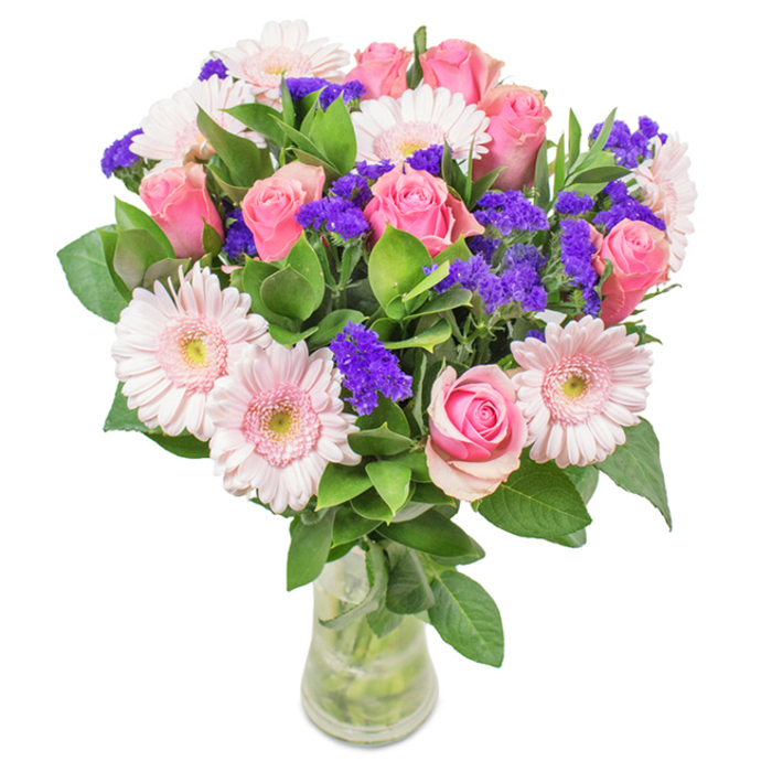 A carefully arranged floral bouquet sits in a crystal-clear vase atop a light surface, illuminated by soft daylight-crafted by expert florists from Flowers Chadwell Heath. The bouquet's heart lies in its approximately eight velvety pink roses, many just beginning to unfurl their petals into perfect spirals, nestled closely among six pale pink gerbera daisies noted for their bright, open faces and almost white central disks. Between these, vivid purple statice flowers add texture and depth with their clustered, papery blossoms, creating lively visual contrast alongside the abundance of lush dark green foliage. The greens spill generously from the base, imparting a freshness and fullness so often admired by locals passing the florists near High Road. Every element is precisely placed for maximum harmony-stems exposed below the water line sparkle in the clear vase, hinting at the bouquet's recent freshness and its journey from local flower wholesalers into skilled hands. Perfect for gifting on birthdays, anniversaries, Mother's Day, or to uplift a friend recovering nearby, this composition radiates joy, appreciation, and heartfelt celebration-a distinctive creation that brings the vibrant charm of Chadwell Heath's community right to your door.